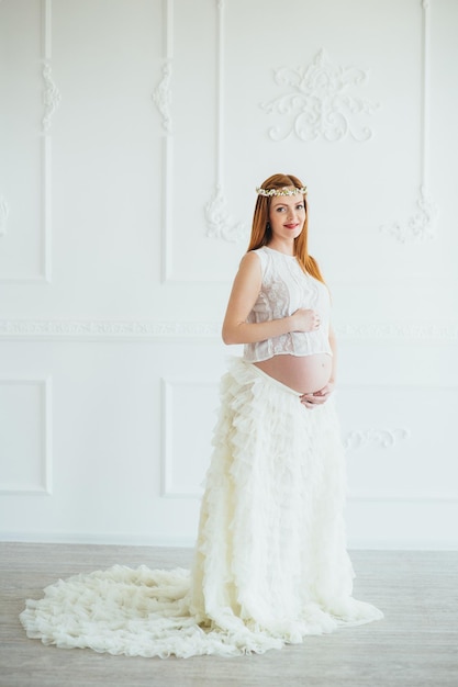 Redhaired pregnant young girl in a white dress near the white wall