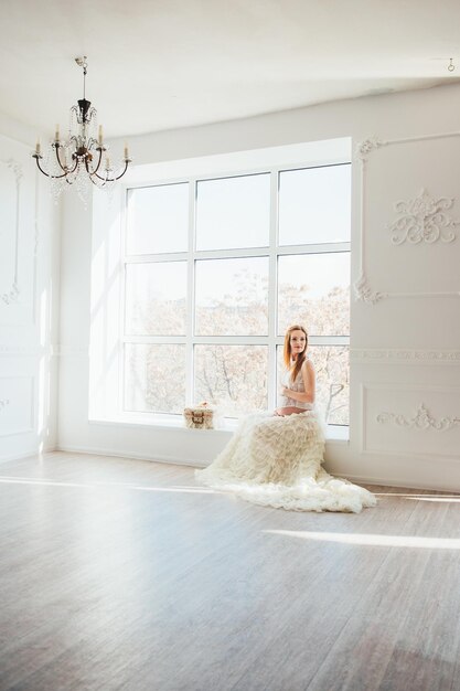 Redhaired pregnant young girl in a white dress near the window