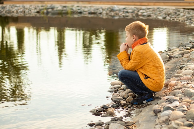 Photo redhaired teenager boy in yellow clothes squatting meditating looking at water of pond in city park