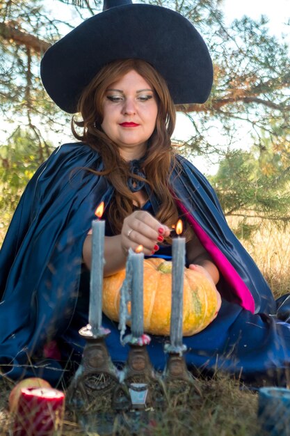 Photo a redhaired witch in a blue cloak and hat in the forest with a pumpkin in her hands conjures candles