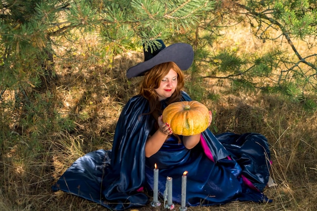 Redhaired witch in a pointed hat and cloak with a pumpkin in the forest next to a candle