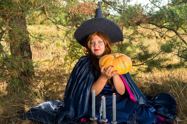 Redhaired witch in a pointed hat and cloak with a pumpkin in the forest