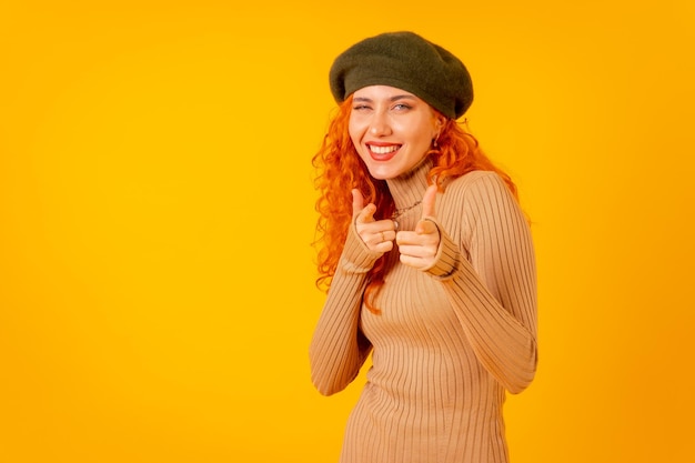 Redhaired woman in beret in studio on a yellow background copy space pointing at camera