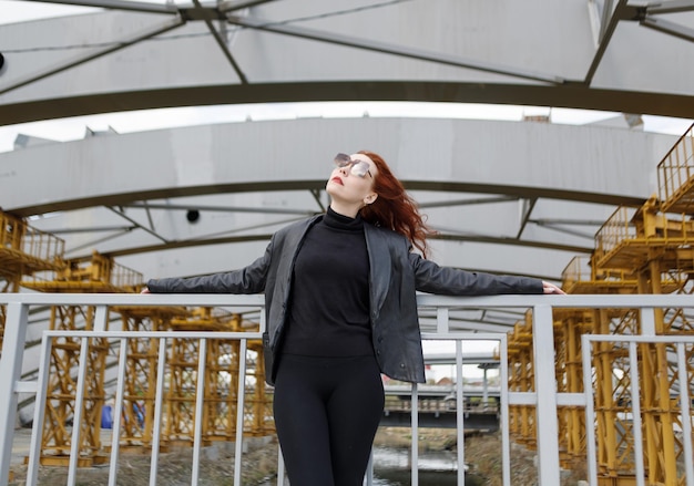 Redhaired woman in glasses and a black leather jacket against the background of an urban landscape