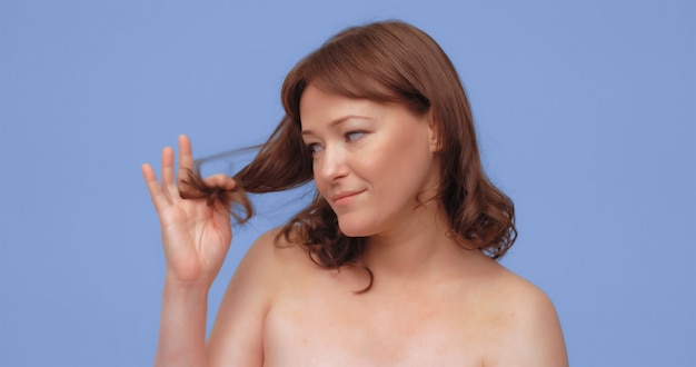 Redhaired woman holds and looks at split ends of her hair adult lady with naked shoilders cut out on...