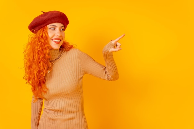 Redhaired woman in a red beret on a yellow background studio shot pointing at copy space