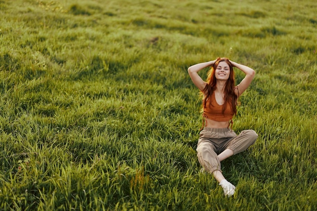 A redhaired woman sits in nature on the grass with her hands behind her head and smiling in the sunset light The concept of healthy rest and meditation for health