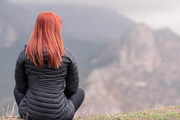 Redhaired woman sitting on top of a cliff observes the mountains out of focus People