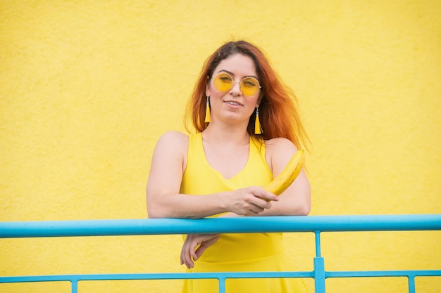 Redhaired woman in sunglasses holds a banana on a yellow background