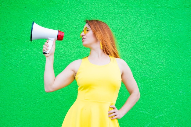 Redhaired woman in sunglasses and a yellow dress holds a megaphone on a green background