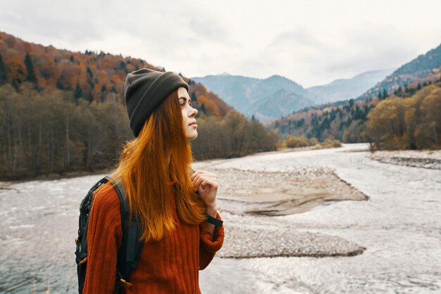 自然の山の川の近くでバックパックと帽子をかぶったセーターを着た赤毛の女性