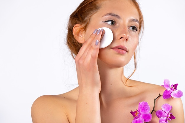 The redhaired woman takes off her makeup with cotton pads\
natural everyday makeup and care concept on white wall