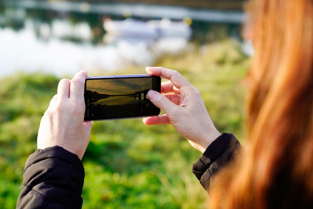 赤い髪の女性は、湖の川の近くの自然の中でスマートフォンで写真を撮ります