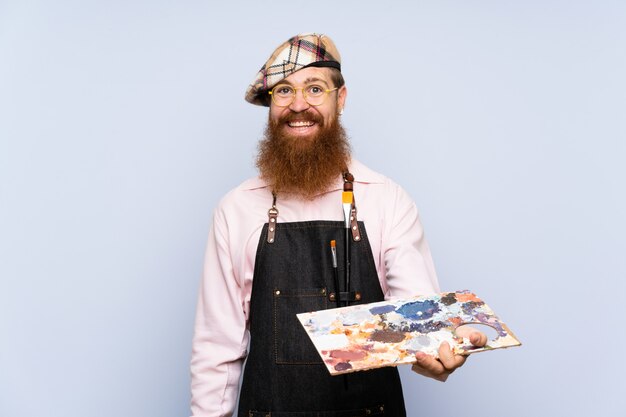 Redhead artist man with long beard holding a palette over isolated blue wall with happy expression