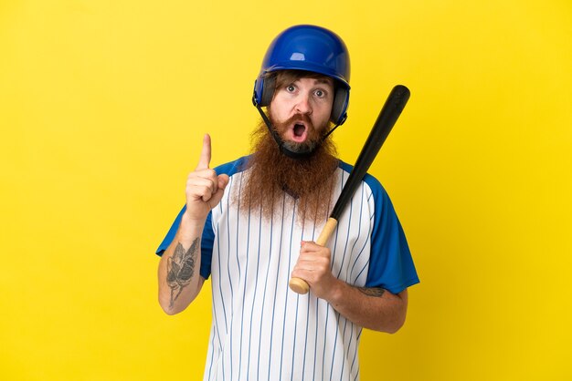 黄色の背景に分離されたヘルメットとバットを持つ赤毛の野球選手の男は、指を持ち上げながら解決策を実現しようとしています