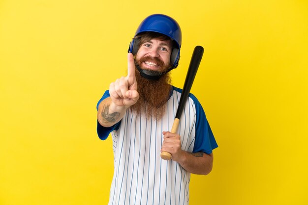 Redhead giocatore di baseball uomo con casco e pipistrello isolato su sfondo giallo che mostra e solleva un dito