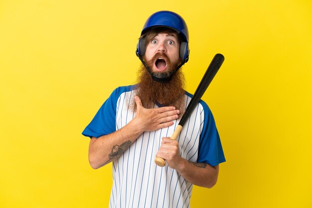 Foto redhead giocatore di baseball uomo con casco e pipistrello isolato su sfondo giallo sorpreso e scioccato mentre guarda a destra