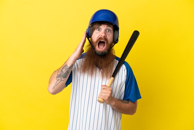 Foto uomo del giocatore di baseball della testarossa con casco e pipistrello isolato su sfondo giallo con espressione facciale sorpresa e scioccata