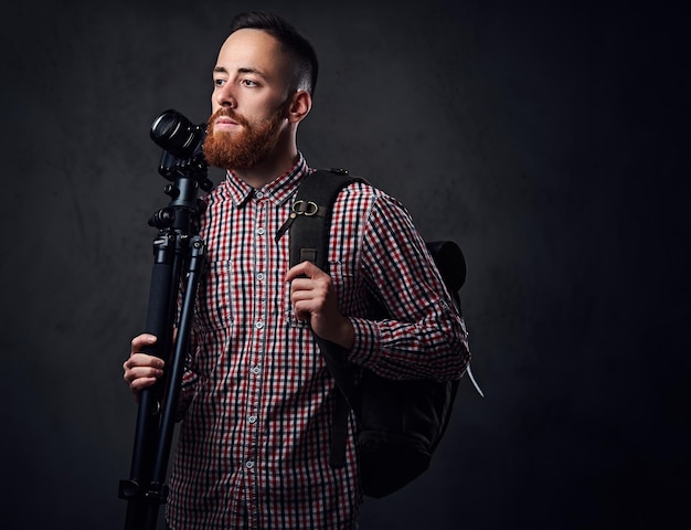 Рыжий бородатый фотограф-фрилансер со штативом и рюкзаком.