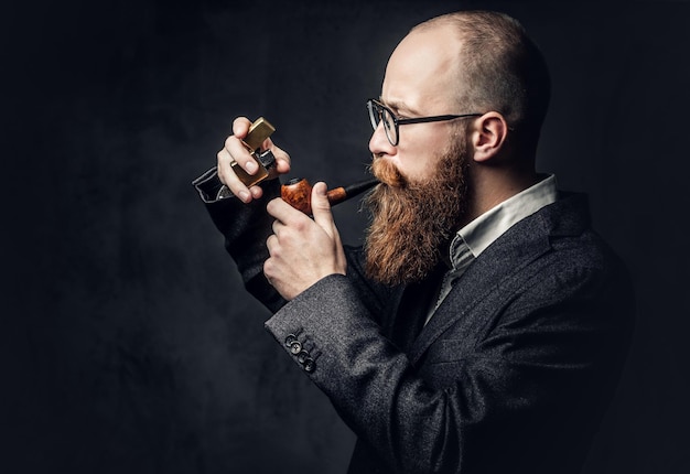 Redhead bearded male dressed in a suit and eyeglasses smoking tradition pipe over dark grey background.