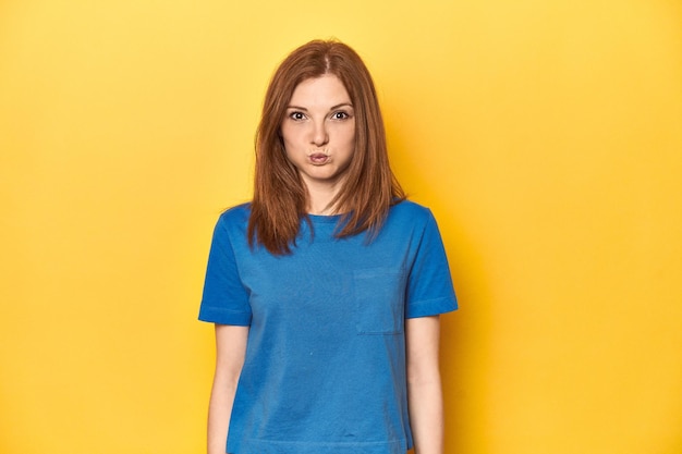 Redhead in blue tshirt on yellow backdrop blows cheeks has tired expression Facial expression