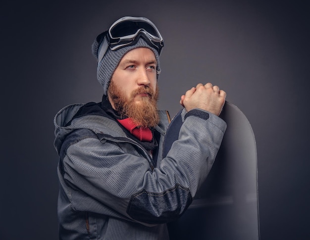 Redhead brutal snowboarder with a full beard in a winter hat and protective glasses dressed in a snowboarding coat posing with snowboard at a studio, looking away. Isolated on a gray background.