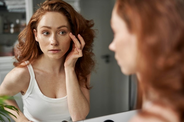 Foto donna caucasica dai capelli rossi nel bagno che controlla la condizione del viso