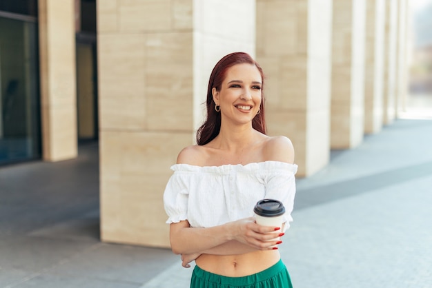 Redhead caucasian woman in her thirties laughs and holds a cup with coffee on the background of a modern building.