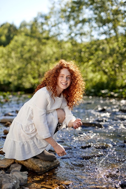 빨간 머리 곱슬한 백인 여성은 숲의 강, 호수 근처에 앉아 물을 튀깁니다. 여름 자연 속에서 여성은 행복하게 웃고 혼자 즐겁게 카메라를 바라보고 있습니다. 초상화. 아름다움, 자연 개념