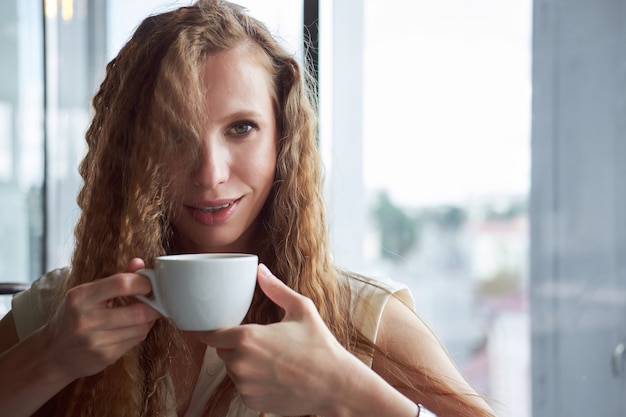 Redhead curly white skinny girl in white shirt drinking coffee and smiling