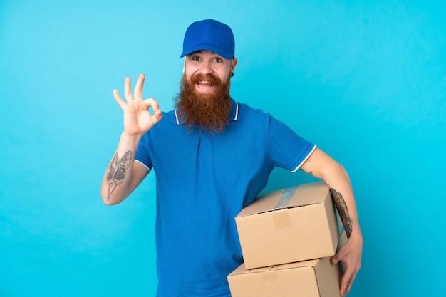 Redhead delivery man over isolated blue wall showing ok sign with fingers