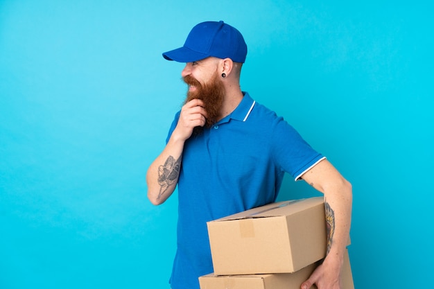 Redhead delivery man over isolated blue wall thinking an idea and looking side