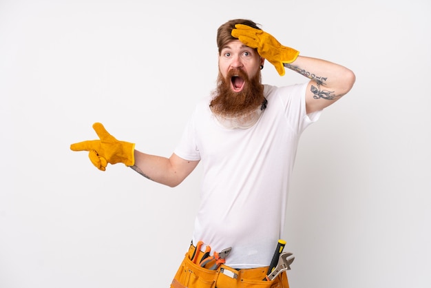 Uomo dell'elettricista di redhead con la barba lunga sopra la parete bianca isolata sorpresa e puntando il dito al lato