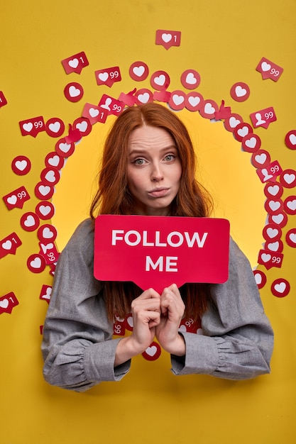 Photo redhead female holding follow me sign, ask to be more active in internet