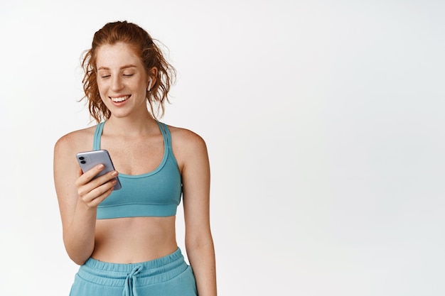 Redhead fitness girl in activewear choosing music in smartphone app while workout in gym wearing wireless headphone standing against white background