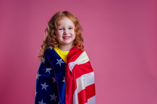 Redhead zenzero affascinante femmina bambino celebrazione indipendenza tenendo bandiera americana su uno sfondo rosa in studioil concetto di apprendimento della lingua inglese e il sogno di libertà