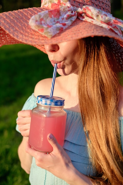 Redhead ginger girl dressed in coral color hat and blue dress drinks pink smoothie from glass mug in summer