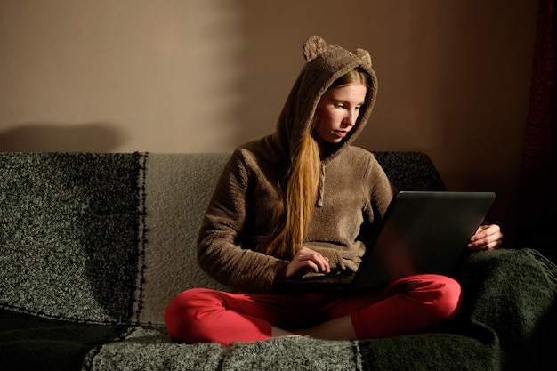 Redhead ginger girl in funny hoody working with laptop, sitting on the sofa