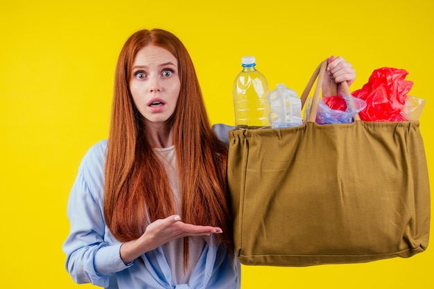 Redhead ginger girl with consternation on her face pointing finger copyspase with bad full of noneco plastic trash in studio yellow background