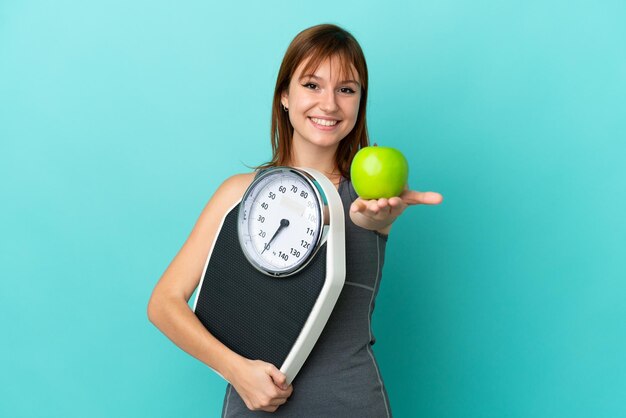 Redhead girl isolated on blue background holding weighing machine and offering an apple