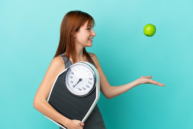 Redhead girl isolated on blue background with weighing machine and with an apple
