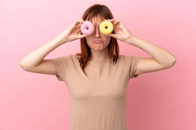 Redhead girl isolated on pink background holding donuts in eyes with sad expression