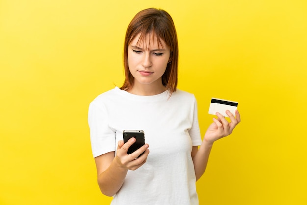 Redhead girl isolated on yellow background buying with the mobile with a credit card