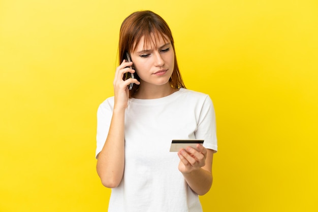 Redhead girl isolated on yellow background buying with the mobile with a credit card