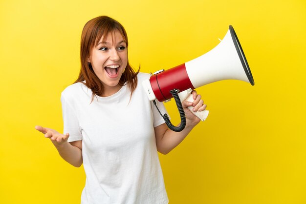 Redhead girl isolated on yellow background holding a megaphone and with surprise facial expression