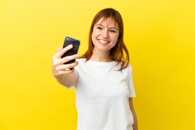 Redhead girl isolated on yellow background making a selfie with mobile phone