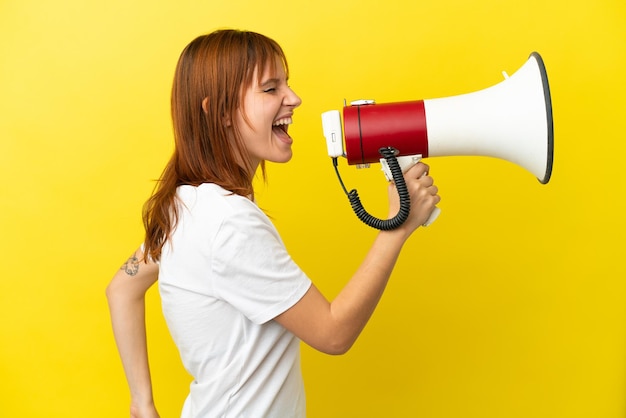 Redhead girl isolated on yellow background shouting through a megaphone to announce something in lateral position