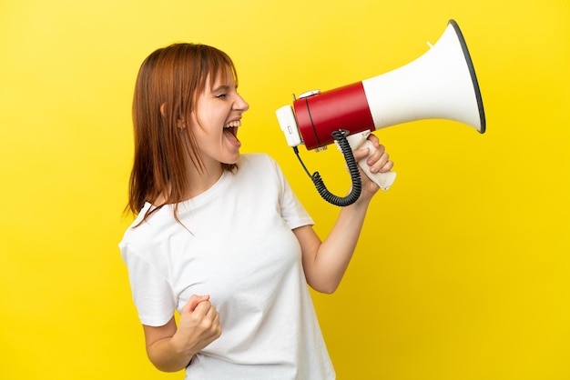 Redhead girl isolated on yellow background shouting through a megaphone to announce something in lateral position