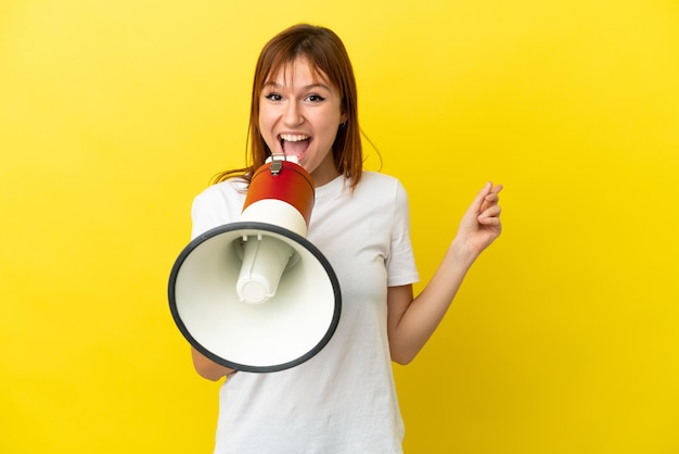 Redhead girl isolated on yellow background shouting through a megaphone and pointing side