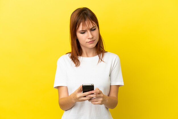 Redhead girl isolated on yellow background using mobile phone